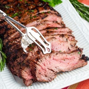 Silver tongs picking up slices of grilled flank steak from a platter.