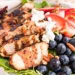 A closeup of a salad topped with sliced chicken, fresh berries, and goat cheese with text overlay that says "Blueberry Balsamic Grilled Chicken Salad".