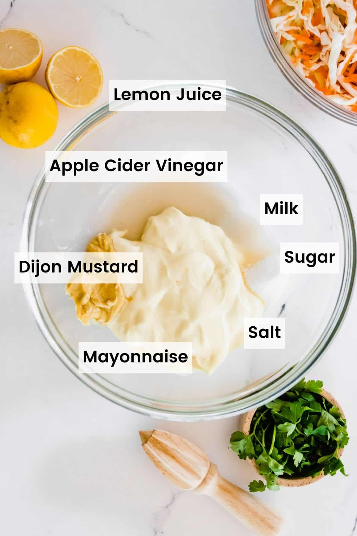 Coleslaw Dressing ingredients in a bowl with text labels including: Lemon Juice, Apple Cider Vinegar, Milk, Dijon Mustard, Sugar, Salt, and Mayonnaise.