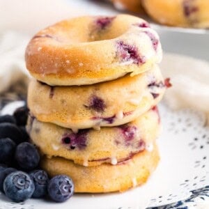 A stack of four blueberry donuts on a white plate with blueberries.