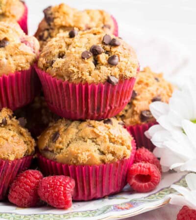 Gluten free raspberry muffins are piled on a plate with fresh raspberries scattered around them.