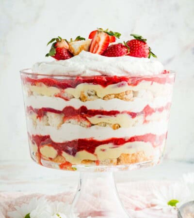 Layers of pastry cream, angel food cake, strawberries, and whipped cream are seen in a glass trifle bowl.