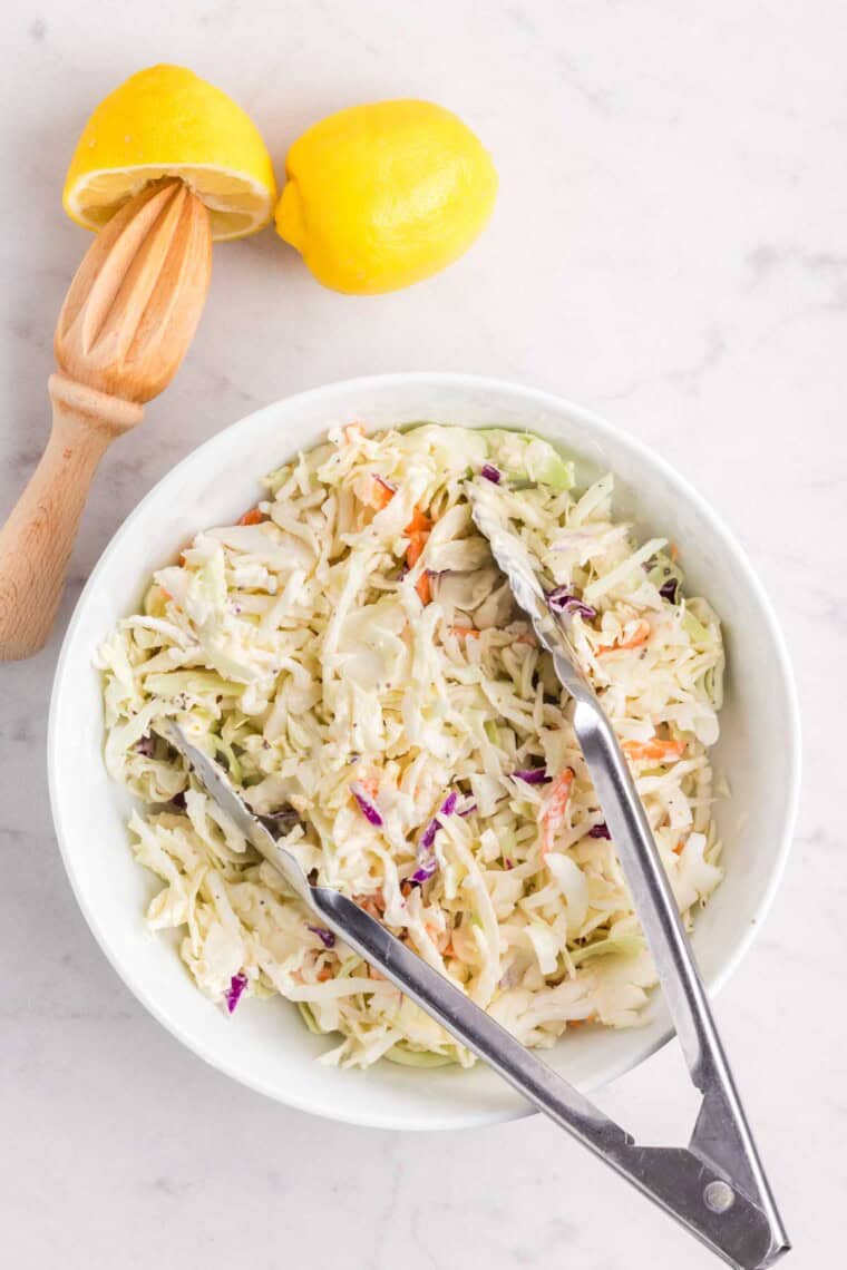 Tongs toss the coleslaw in a bowl.