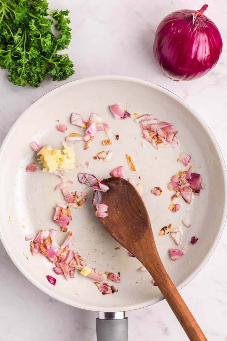 Garlic and red onion are stirred with a wooden spoon as they cook in a white skillet.