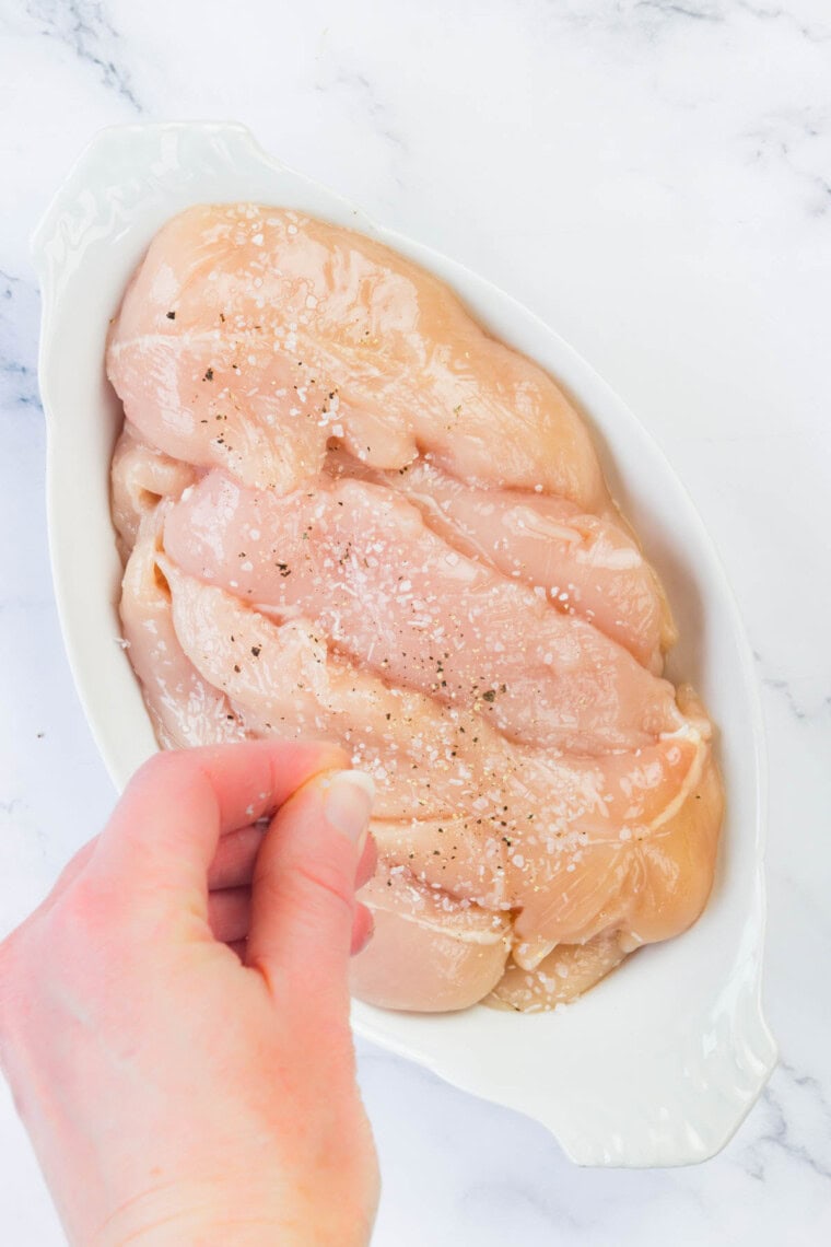 A hand sprinkles salt and pepper into raw chicken tenders.