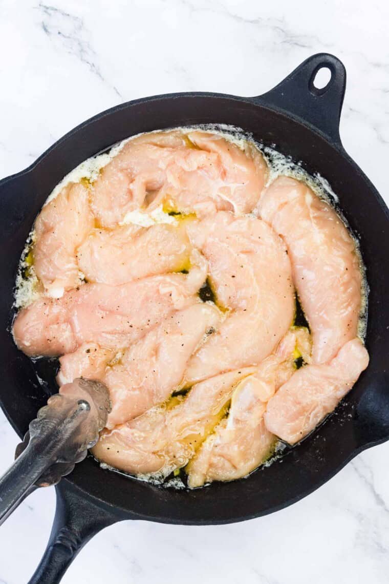 Tongs move and help to cook chicken tenders in a cast iron skillet.