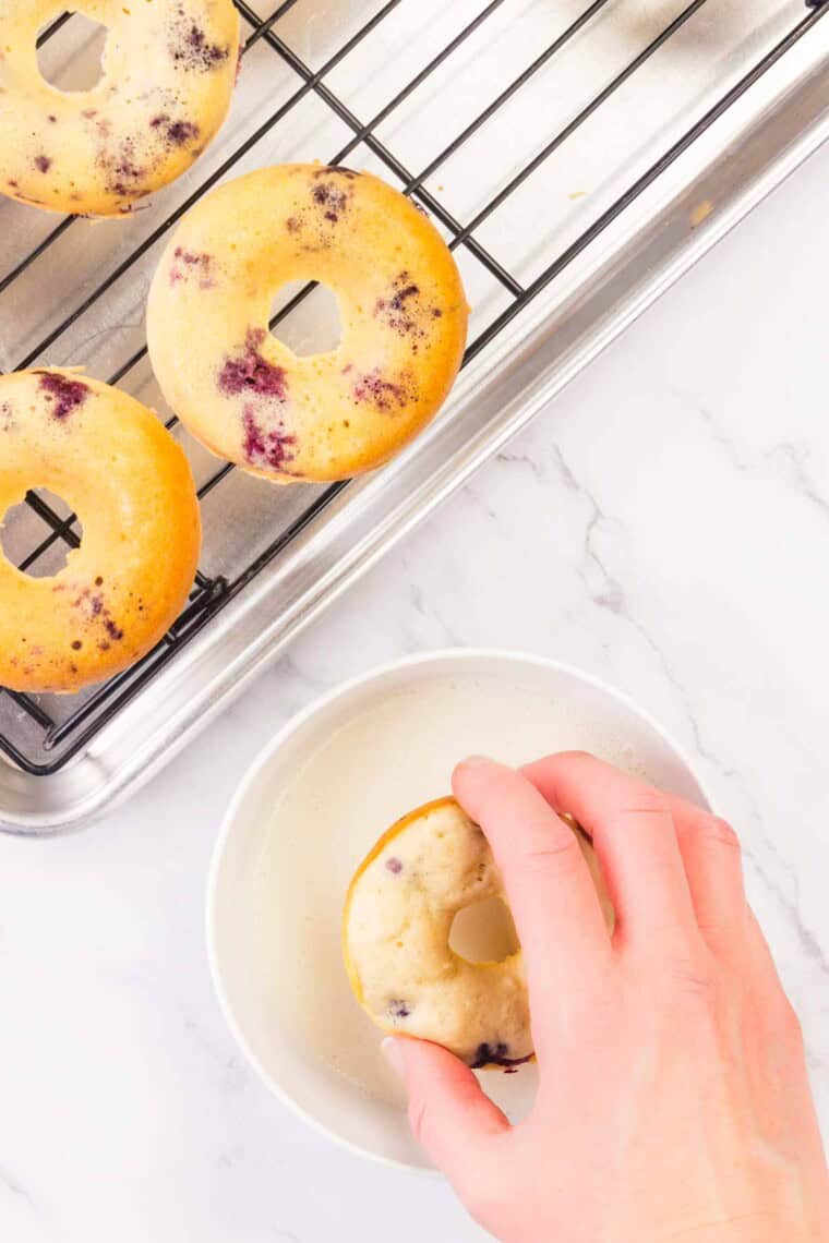 A hand dips a blueberry donut in a bowl of glaze.