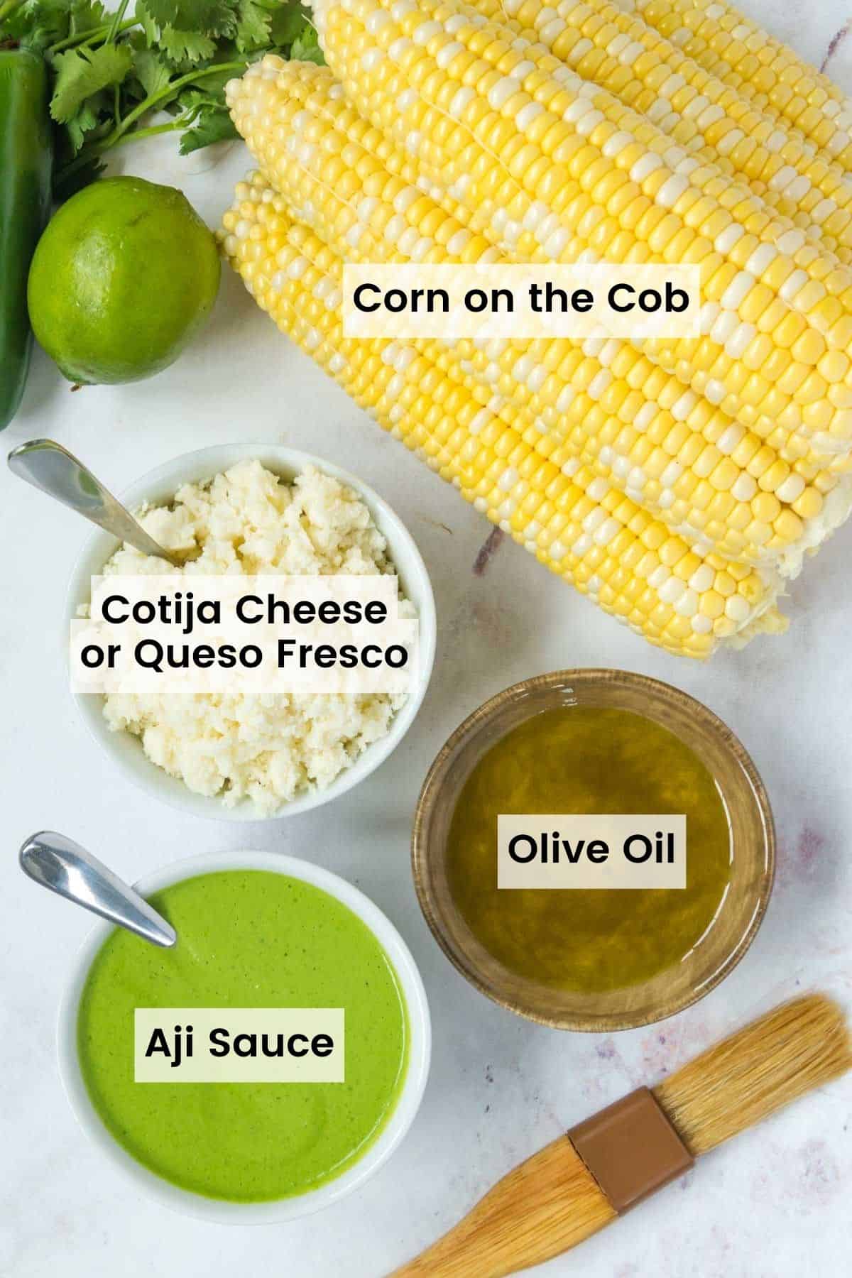 Ingreidents to make peruvian corn on the cob sitting on a marble countertop with text labels, including: corn on the cob, aji sauce, cotija cheese or queso fresco, and olive oil.