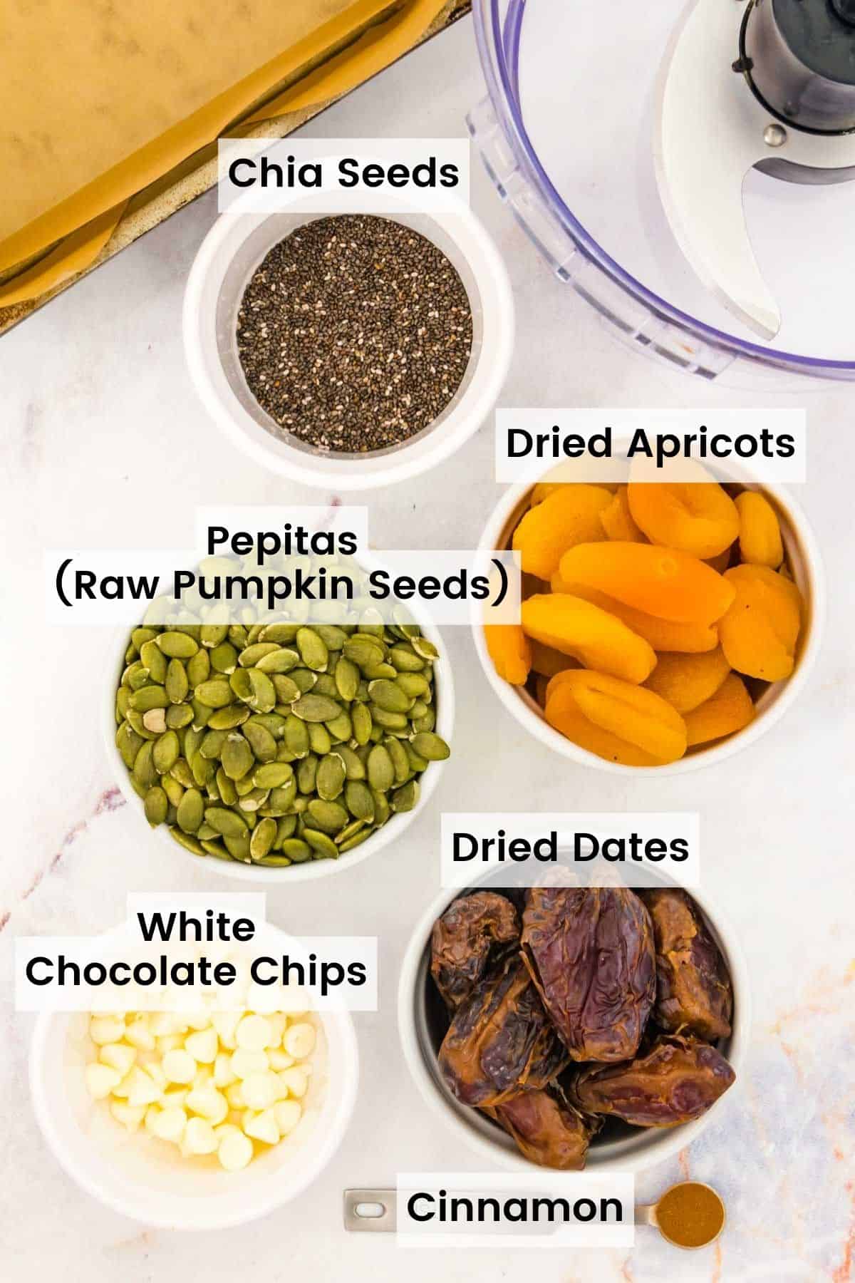 Bowls of ingredients to make energy bars with text labels including: Chia Seeds, Dries Apricots, Pepitas, Dried Dates, White Chocolate Chips, and Cinnamon.