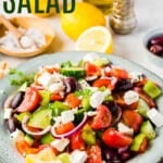 A bowl of cucumbers, tomatoes, olives, red onion, green peppers, and feta tossed in dressing with text overlay that says "Greek Salad".