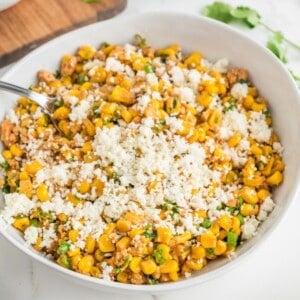 A white serving bowl holds street corn salad with a serving spoon.