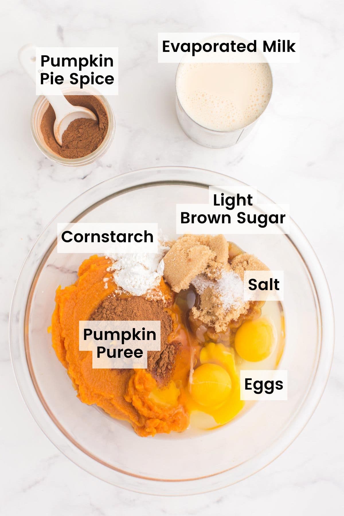 A glass bowl with pumpkin puree, two eggs, light brow sugar, salt, cornstarch, and pumpkin pie spice on a marble counter top with a jar of pumpkin pie spice and a can of evaporated milk with text labels on each ingredient.