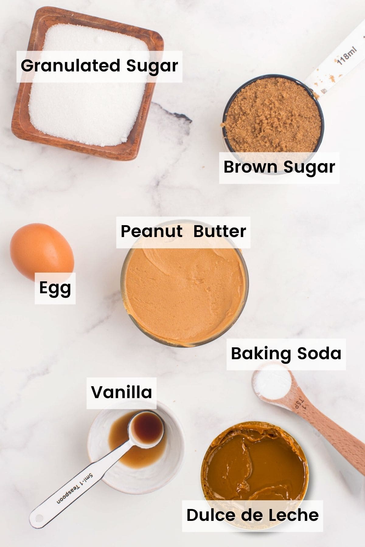 Ingredients to make dulce de leche peanut butter cookies with text labels on a countertop including: granulated sugar, brown sugar, egg, peanut butter, baking soda, vanilla, and dulce de leche.