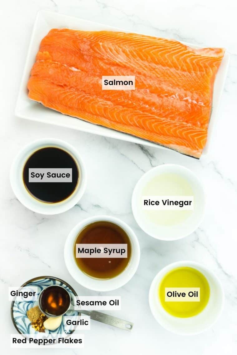 Ingredients to make maple glazed salmon on a countertop with text labels including salmon, soy sauce, rice vinegar, maple syrup, olive oil, sesame oil, garlic, ginger, and red pepper flakes.