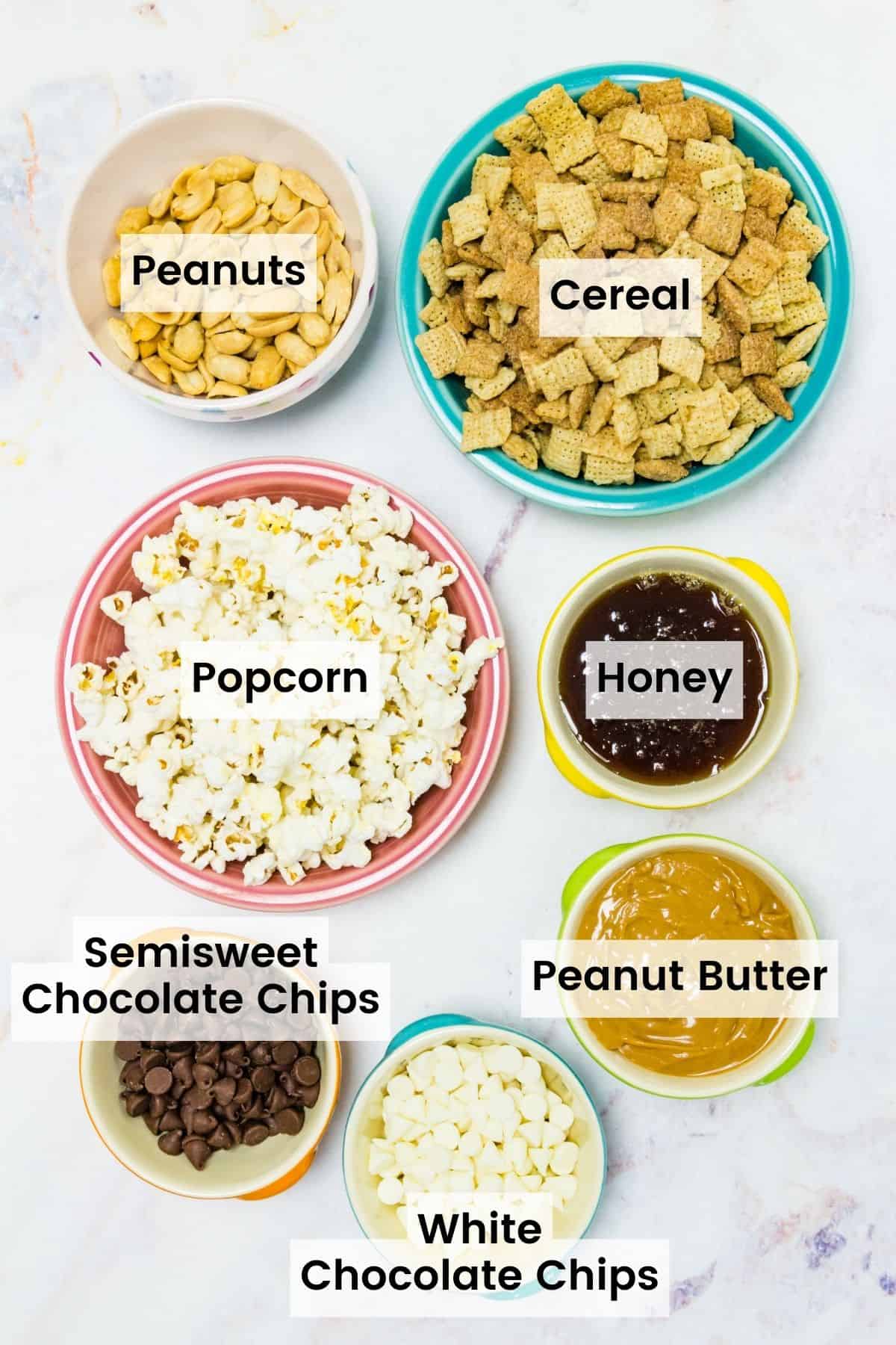 The ingredients for white chocolate popcorn are shown and labeled: popcorn, peanuts, peanut butter, honey, white chocolate chips, semisweet chocolate chips, and cereal.