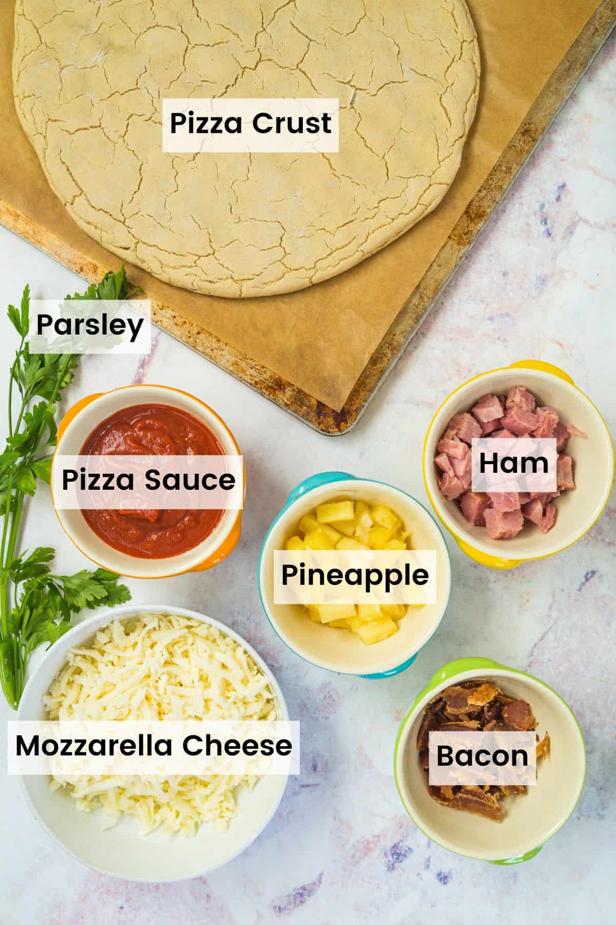 Ingredients to make Hawaiian pizza with text labels including pizza crust on a baking pan with bowls of tomato sauce, cheese, pineapple, cubed ham, and crumbled bacon.