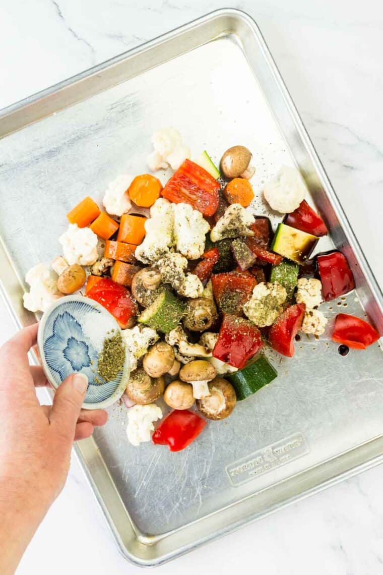 A hand sprinkling seasonings over vegetables on a metal baking sheet.