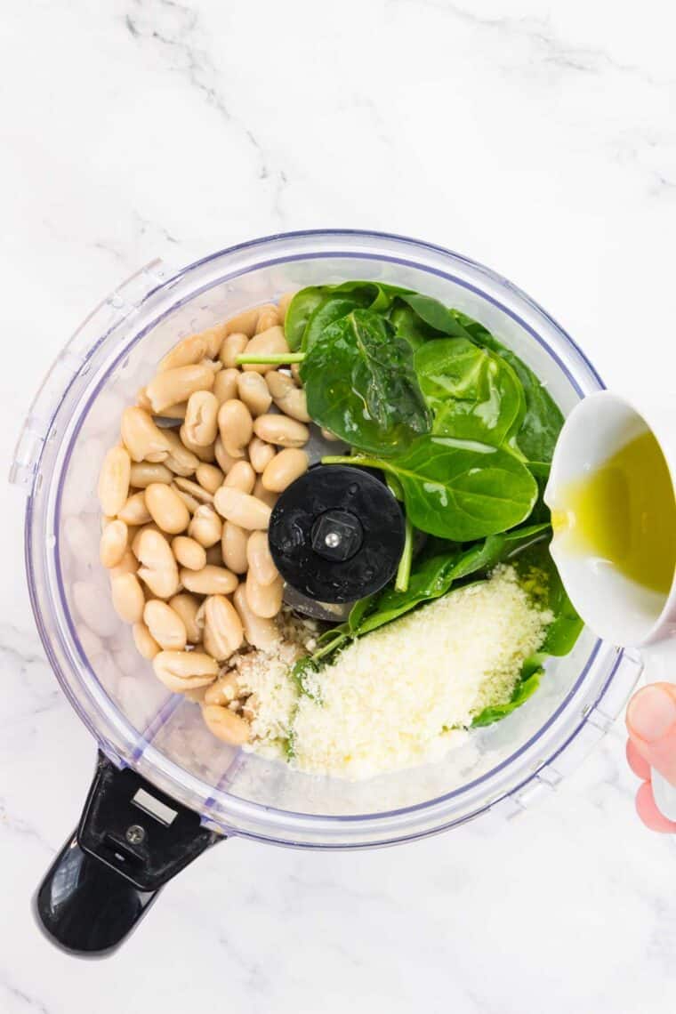 Olive oil is poured into a food processor with spinach, cheese, and white beans.