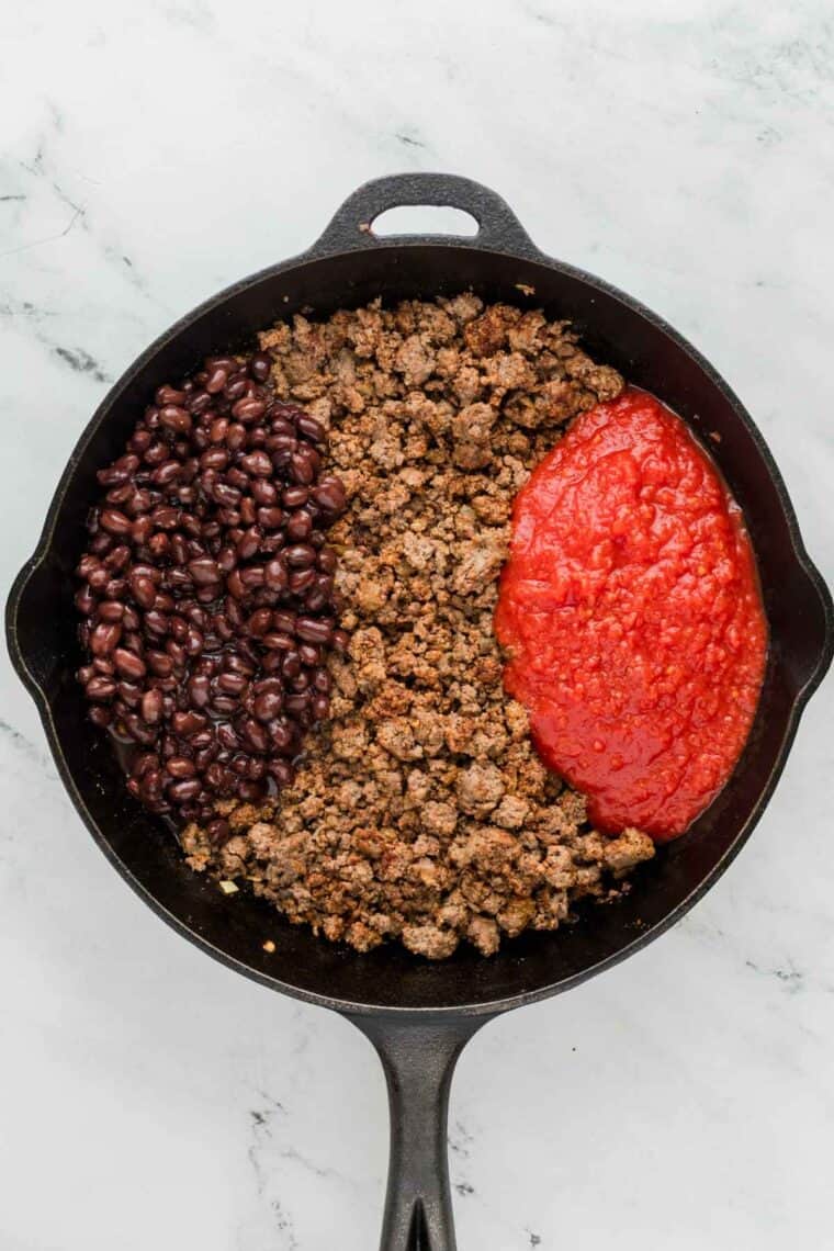 A cast iron pan with three ingredients from left to right: black beans, ground beef, crushed tomatoes.