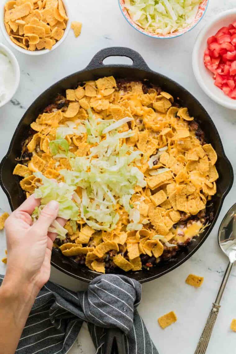 A hand adds shredded cheese to the pan of casserole.