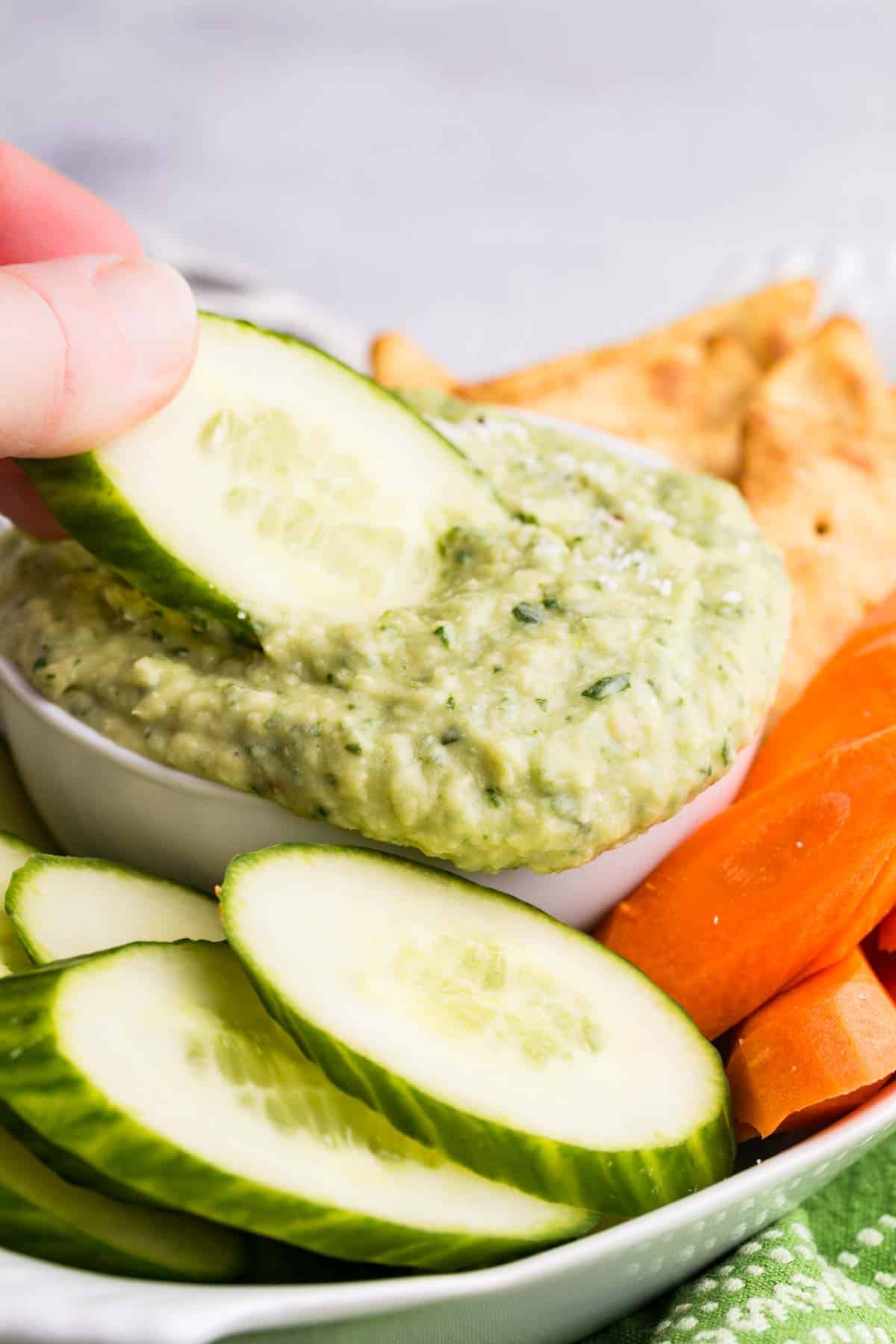 A slice of cucumber is dipped in a bowl of parmesan white bean dip.