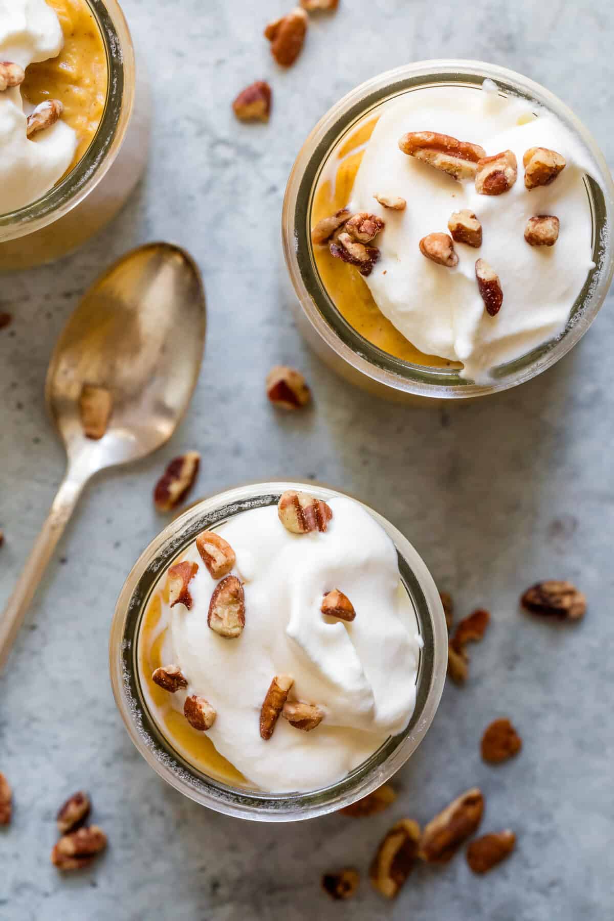 Whipped cream and nuts top two glasses of pumpkin pudding with a spoon to the left.