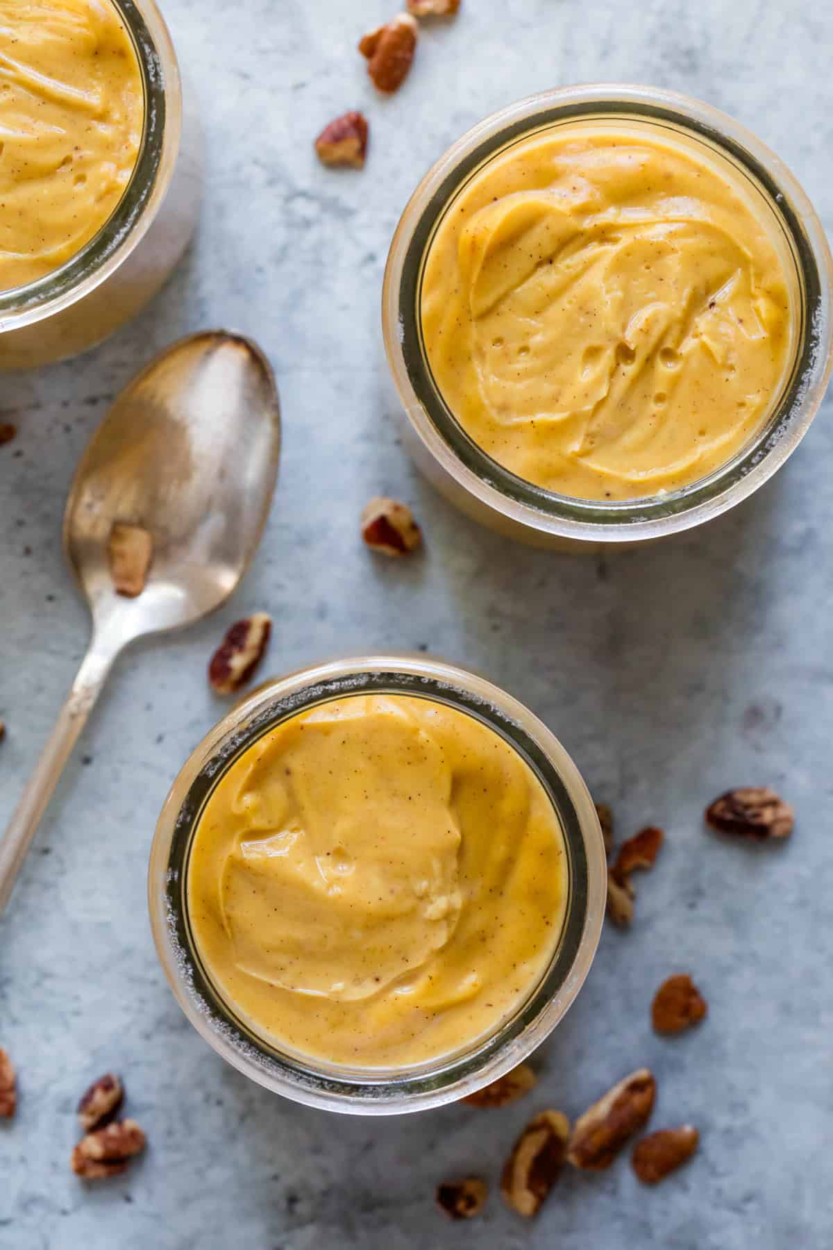 Glass jars of pumpkin pudding with nuts around them and a spoon.