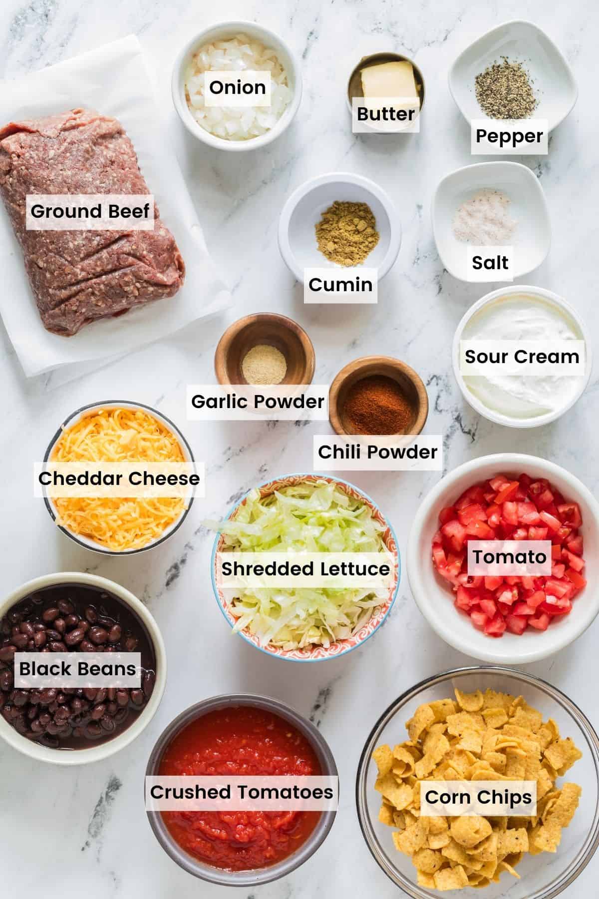 The labeled ingredients for walking taco casserole: ground beef, cheese, crushed tomatoes, corn chips, shredded lettuce, sour cream, black beans, onion.