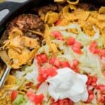 Corn chips are mixed with ground beef and topped with cheese, tomatoes, lettuce, and sour cream in a pan of walking taco casserole.