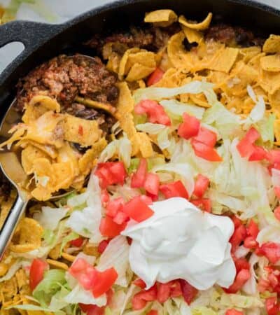 Corn chips are mixed with ground beef and topped with cheese, tomatoes, lettuce, and sour cream in a pan of walking taco casserole.