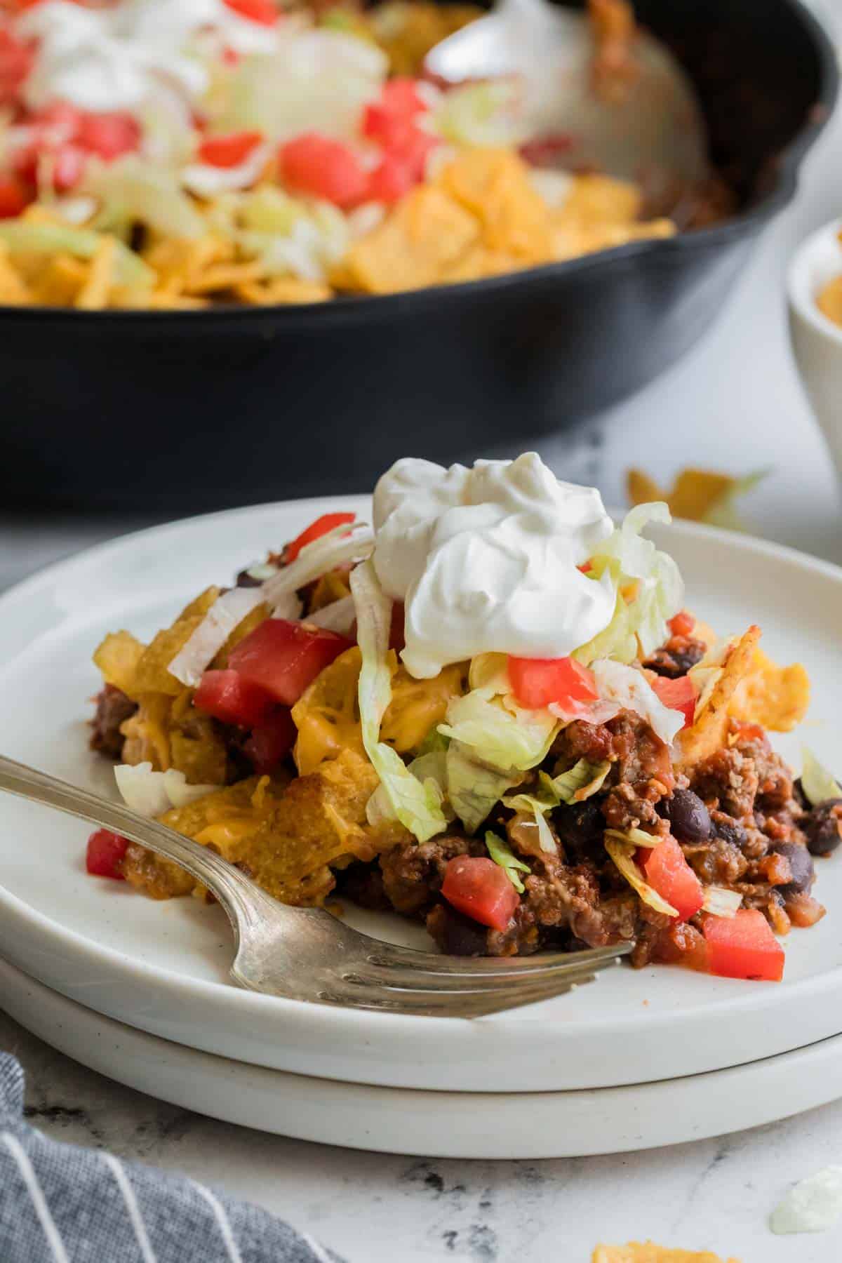 Ground beef spills out of a portion of walking taco casserole topped with sour cream, lettuce, and chopped tomatoes.