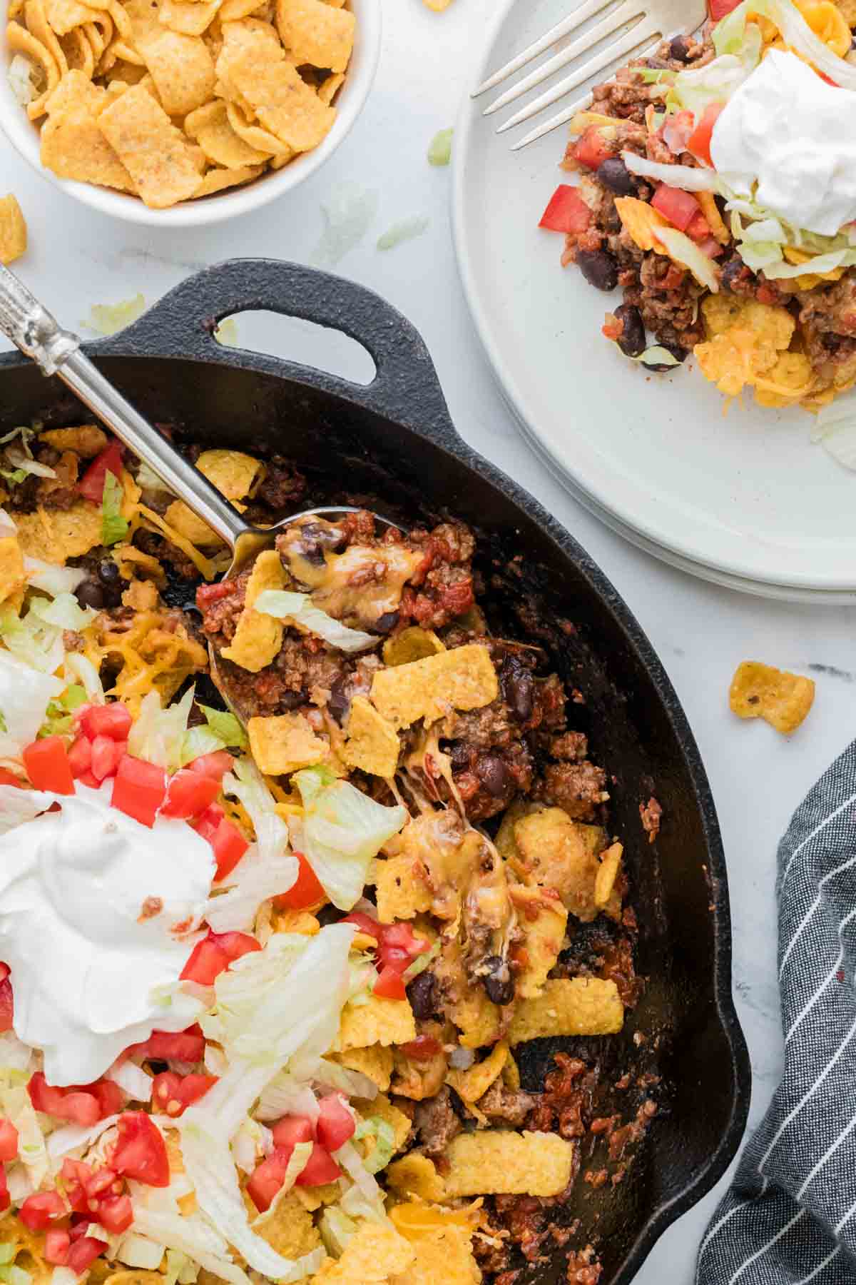 A cast iron pan with walking taco casserole with a plate of casserole to the right.