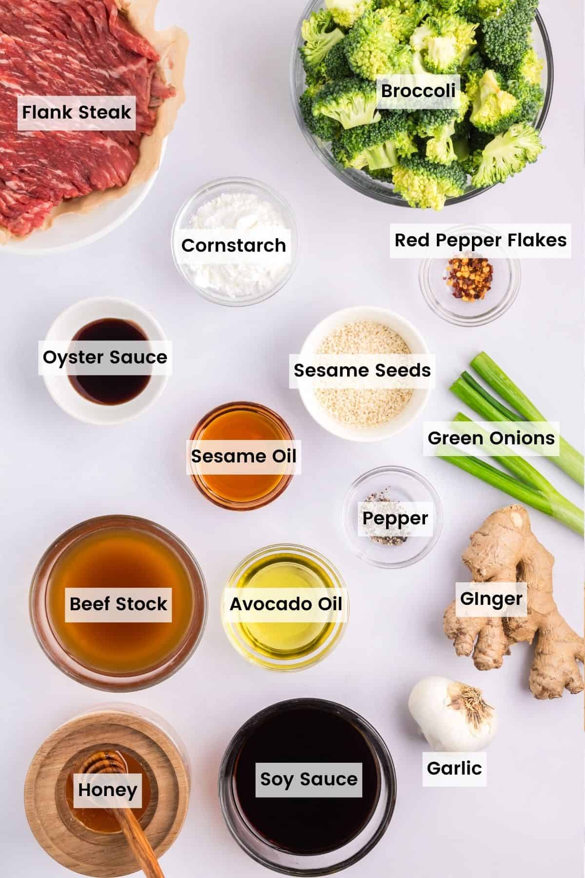 Ingredients to make beef and broccoli portioned out with text labels including flank steak, broccoli, cornstarch, red pepper flakes, oyster sauce, sesame seeds, sesame oil, green onions, pepper, ginger, garlic, beef stock, avocado oil, honey, and soy sauce.