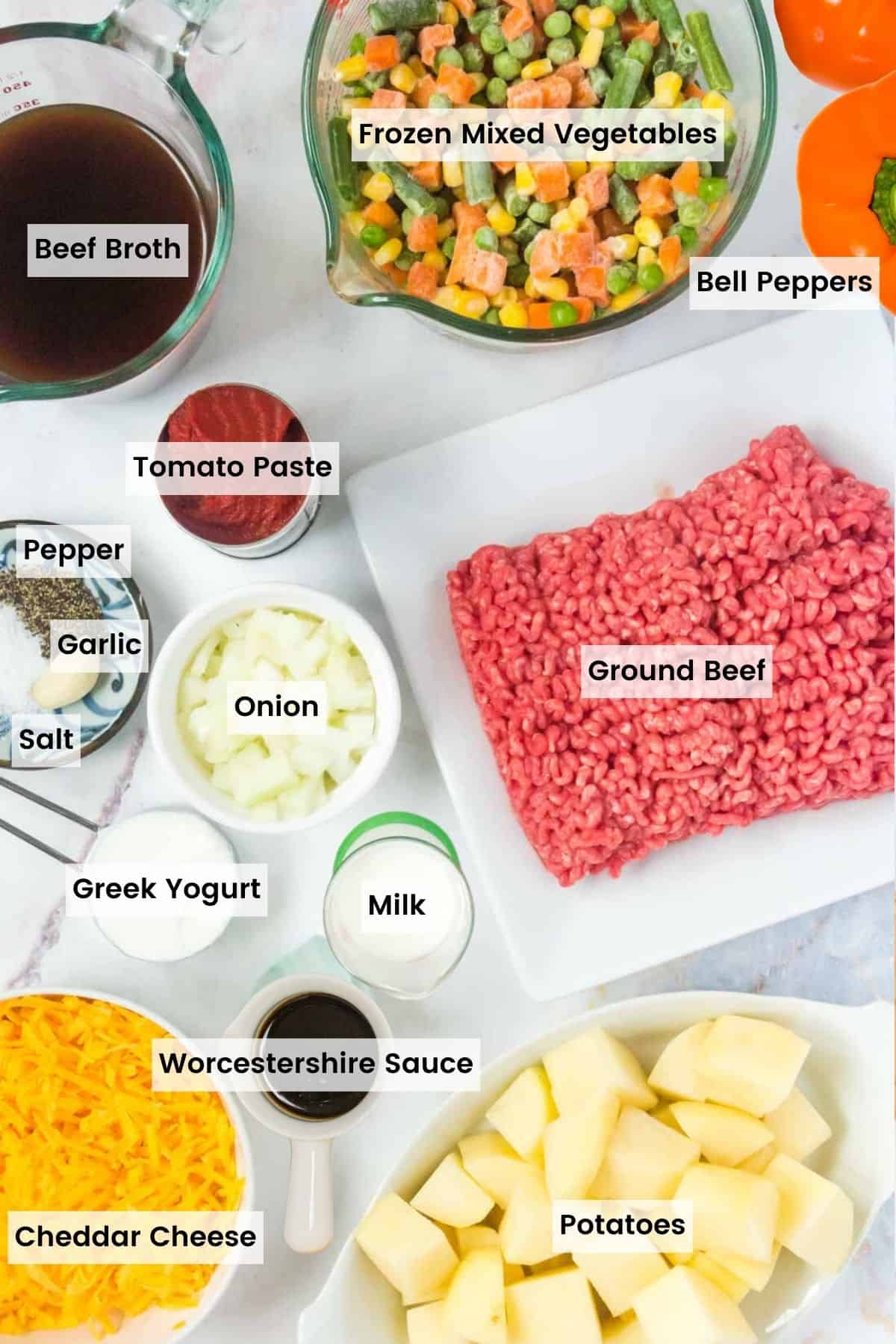 Ingredients to make stuffed peppers portioned out with text labels including: beef broth, frozen mixed vegetables, bell peppers, tomato paste, salt, pepper, garlic, onion, ground beef, milk, Greek yogurt, worcestershire sauce, cheddar cheese, and potatoes.