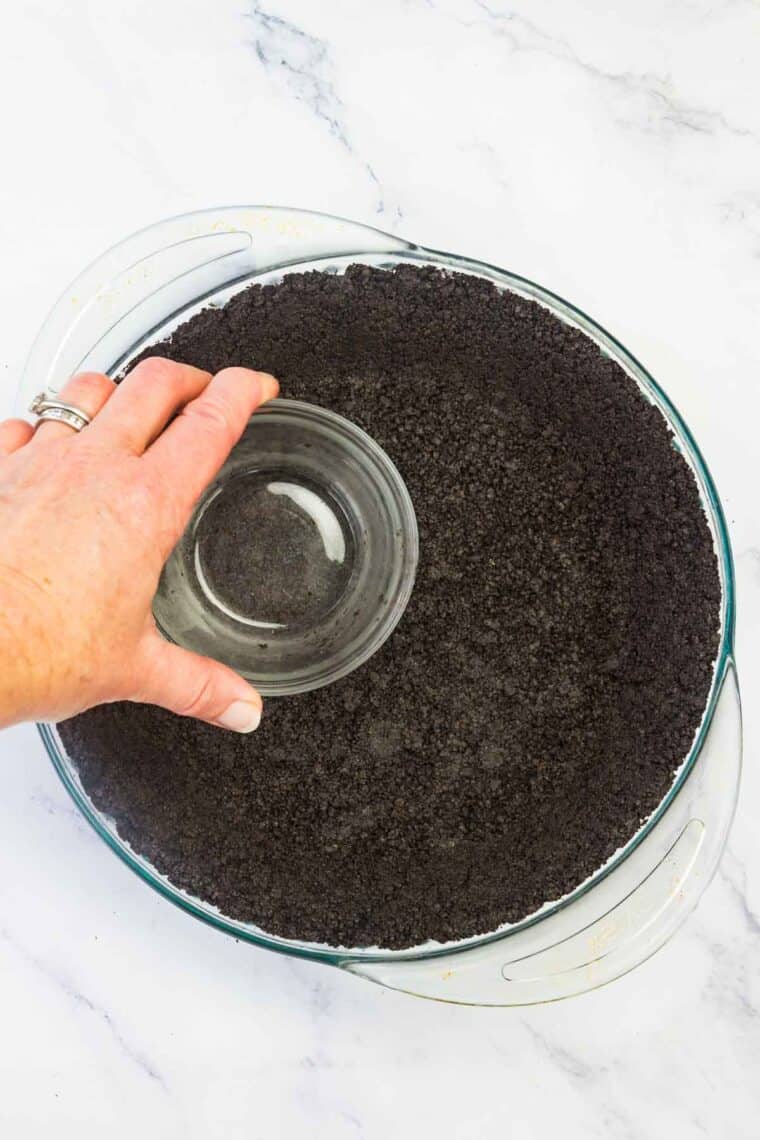 A hand using a glass to press oreo crust into a pie pan.