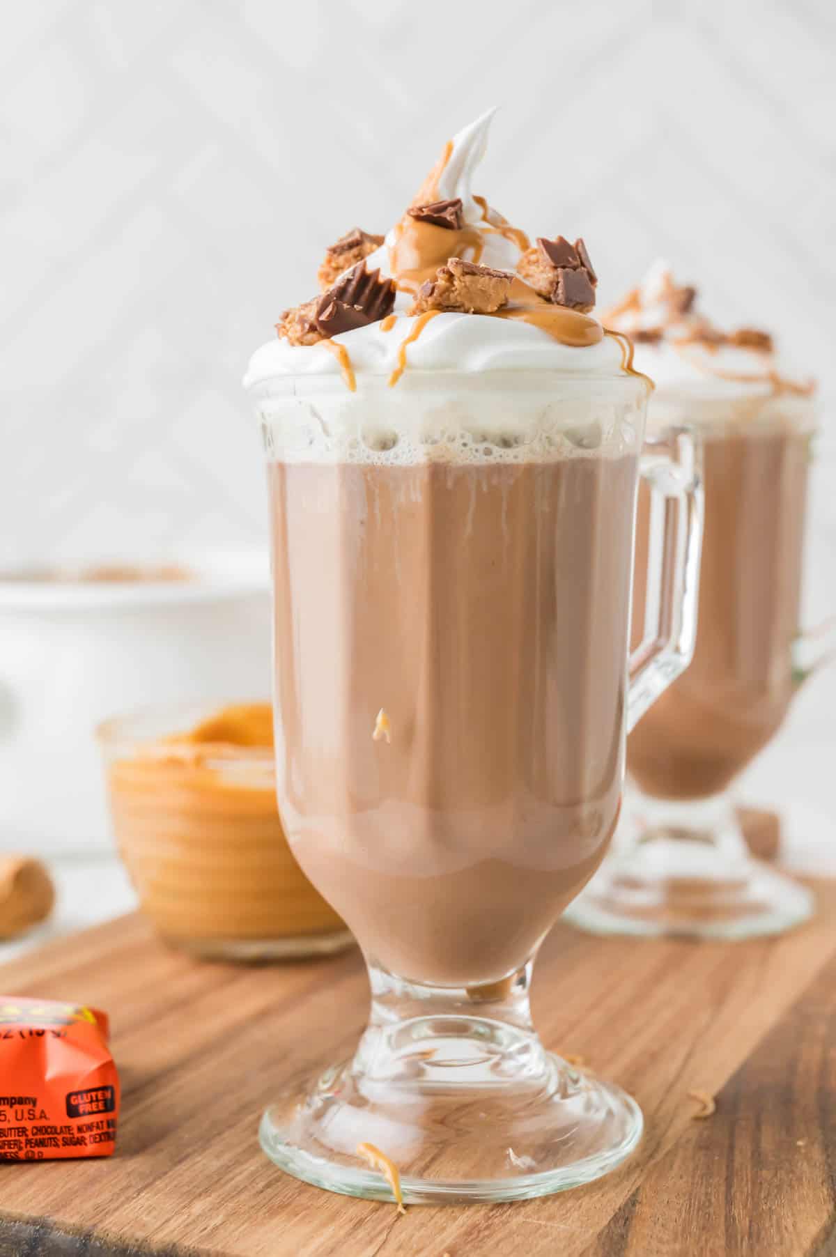 A bowl of peanut butter sauce behind a glass of peanut butter hot chocolate.