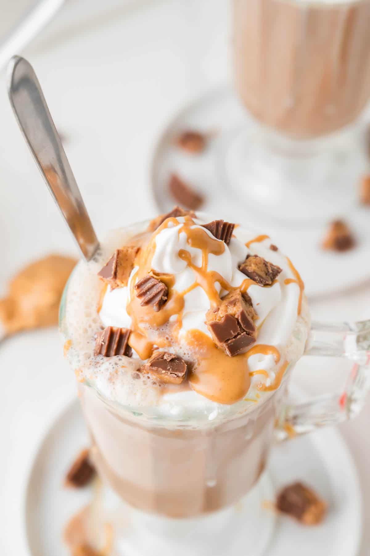 A spoon sticks out of a tall glass of peanut butter hot chocolate topped with whipped cream and reese's.