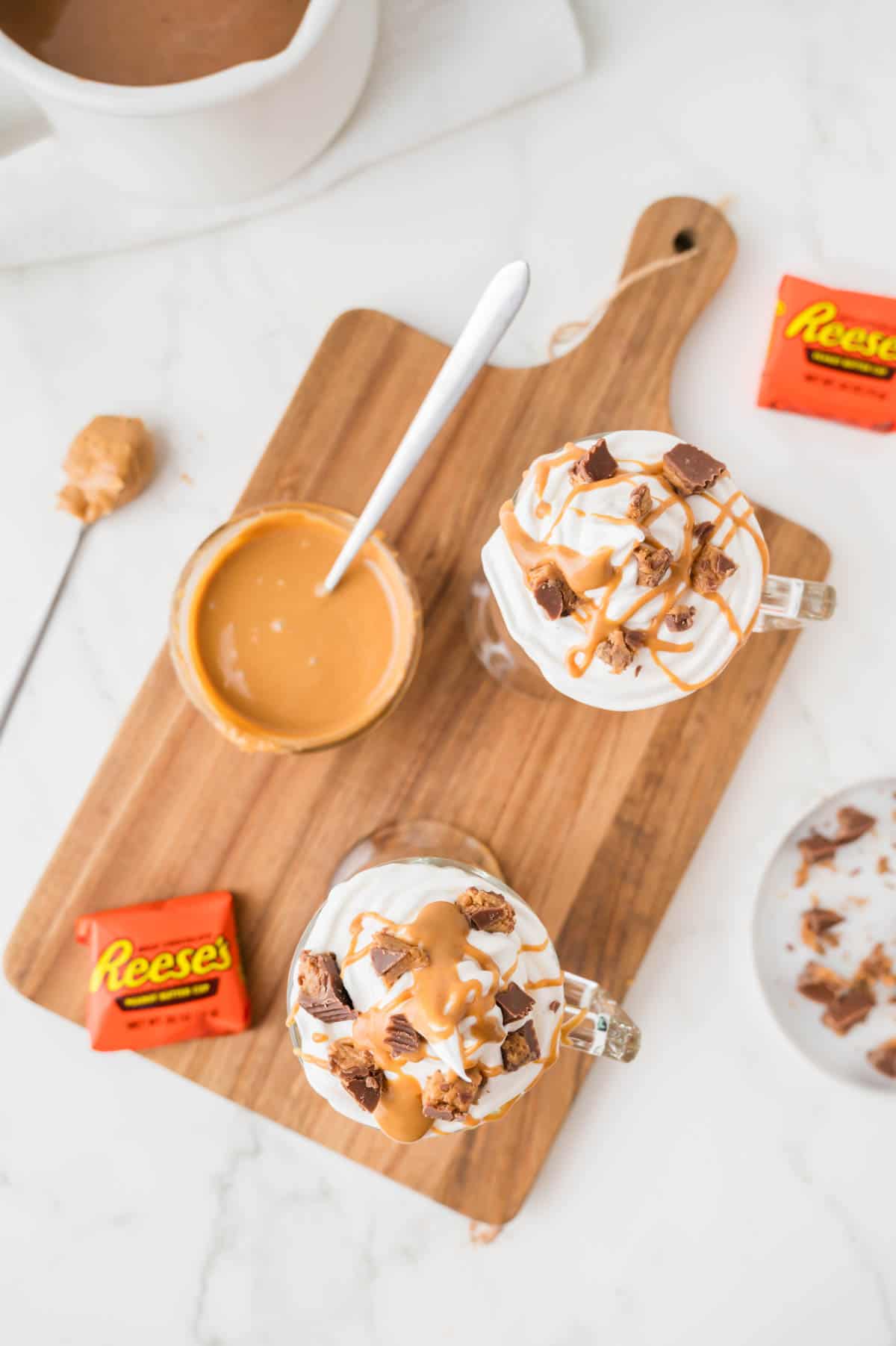 A wooden cutting board holds glasses of peanut butter hot chocolate, reese's peanut butter cups, and peanut sauce.