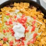 Looking down at a skillet of taco meat topped with cheese, corn chips, and more with text overlay that says "Walking Taco Casserole".