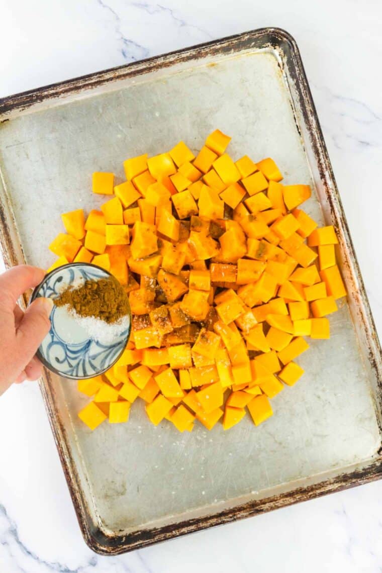 Sprinkling spices over cubes of squash on a sheet pan.