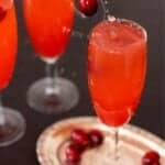 A cranberry being dropped into a flute glass filled with a red drink with text overlay that says "Sparkling Cranberry Mocktail".