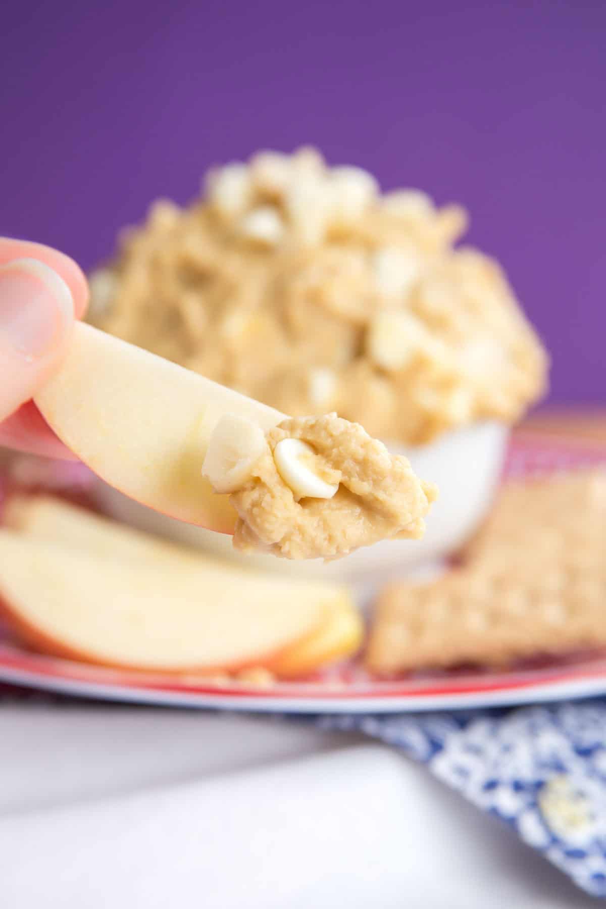 An apple slice dipped in white chocolate macadamia nut cookie dough dip is held up.