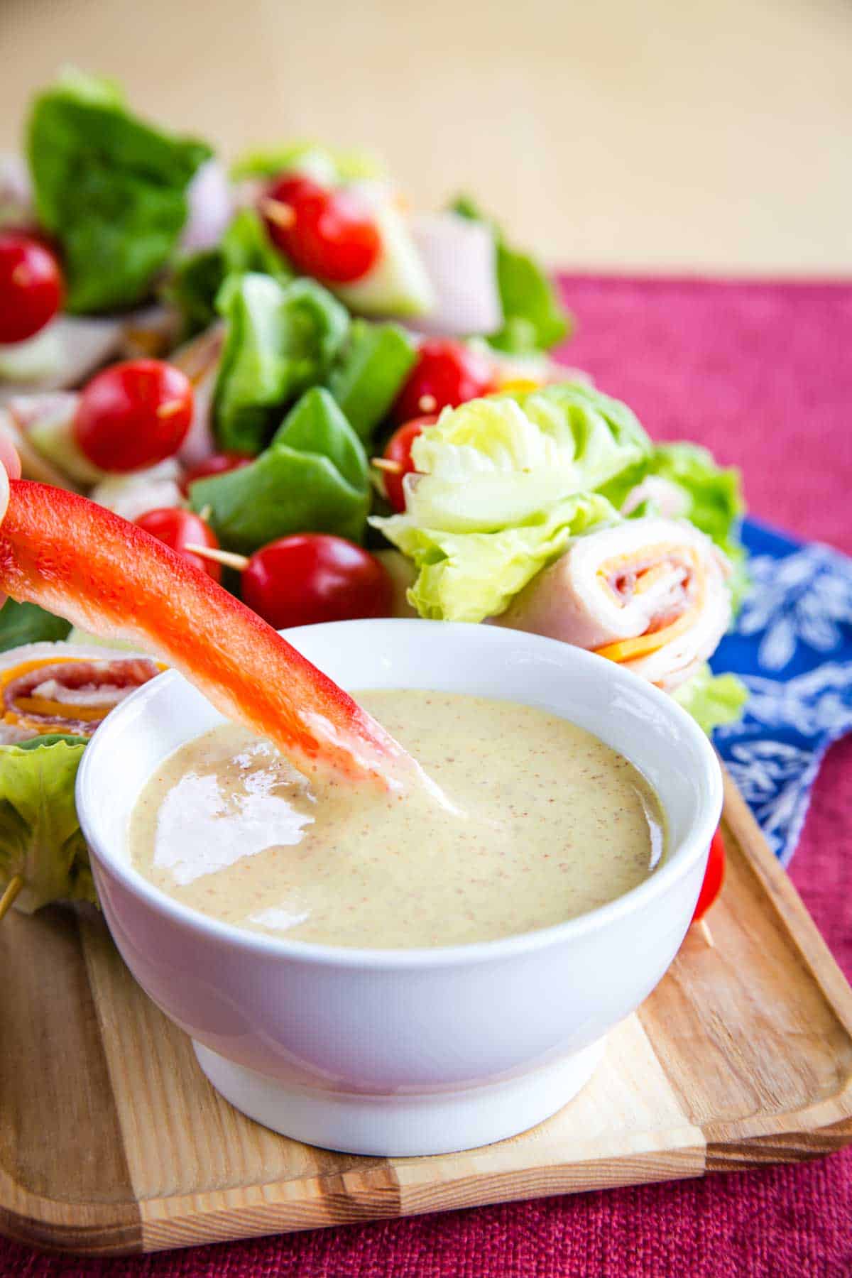 A red pepper slice dipped in a honey mustard dipping sauce with a tray of fresh veggies behind.