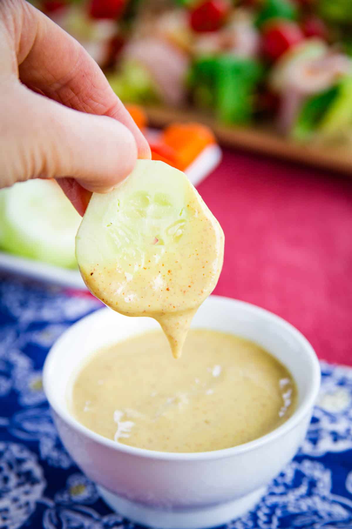 A hand dips a slice of cucumber in a bowl of honey mustard dipping sauce.
