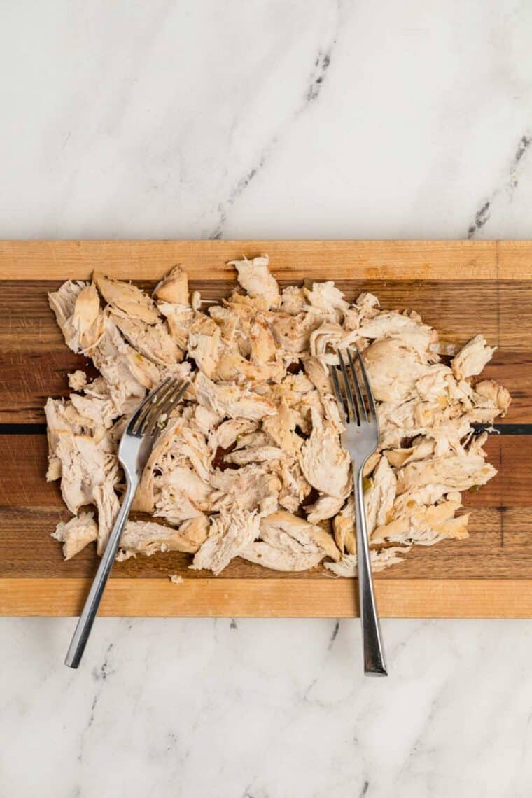 Cooked chicken breast is shredded on a cutting board.