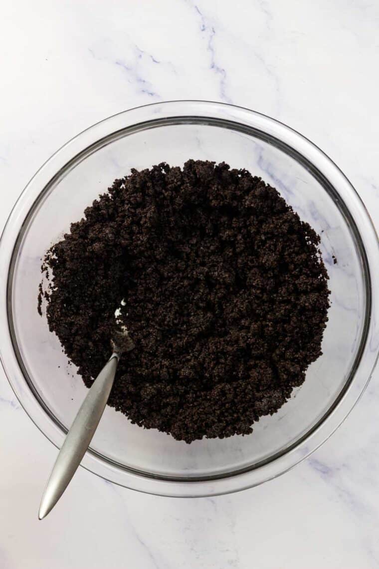 A spoon stirs oreo cookie crumbs and melted butter in a bowl.