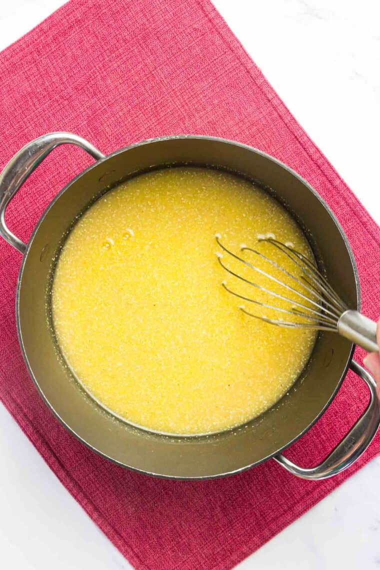 A whisk in a pot of uncooked polenta in chicken broth.