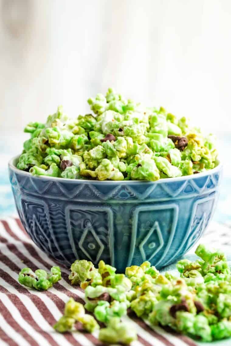 Mint green popcorn is reminiscent of a classic ice cream flavor with homemade popcorn, a little mint extract, and plenty of chocolate chips. A blue bowl filled with green popcorn and more popcorn scattered around it.
