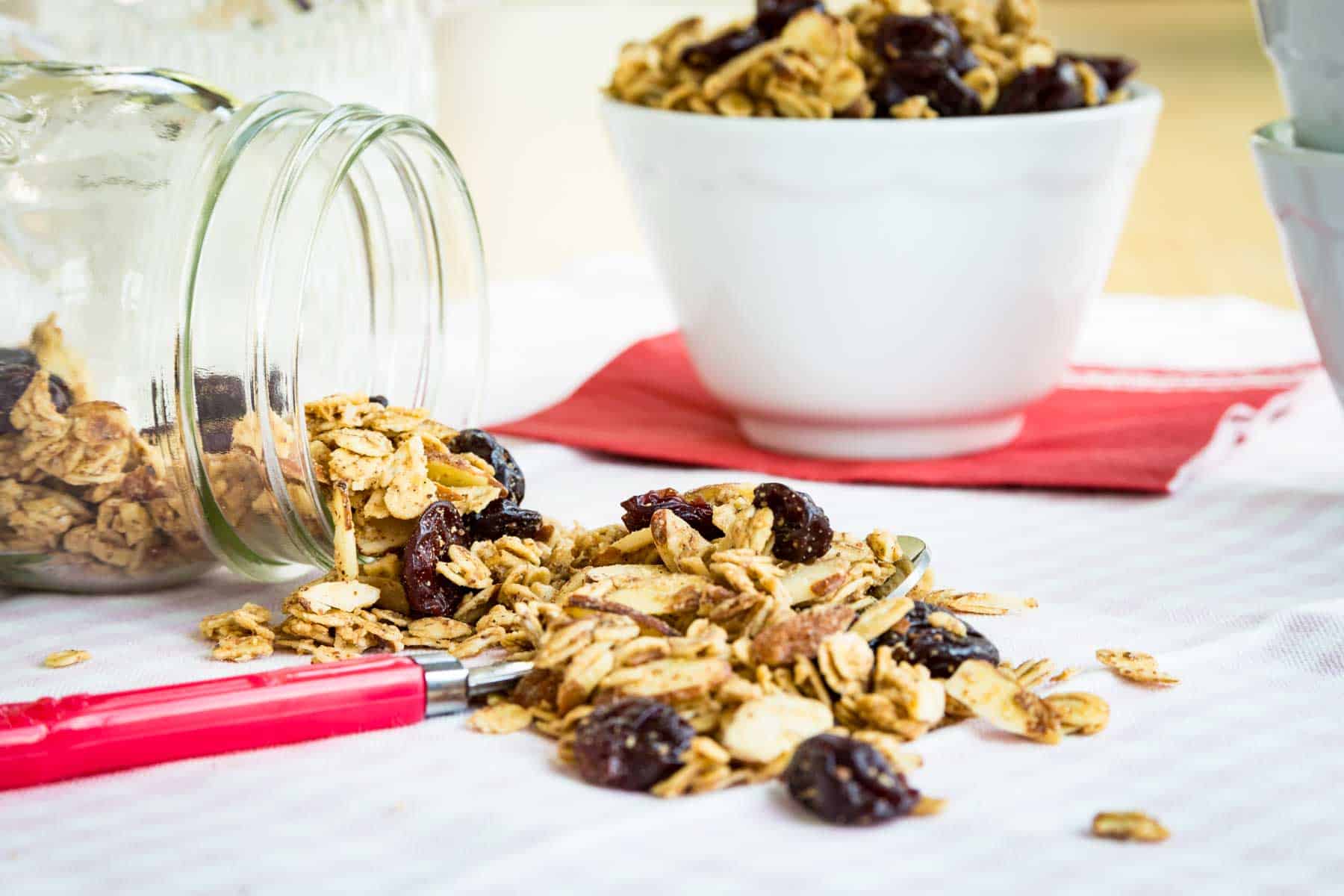 Almond granola spills out of a jar with a bowl of granola behind it.