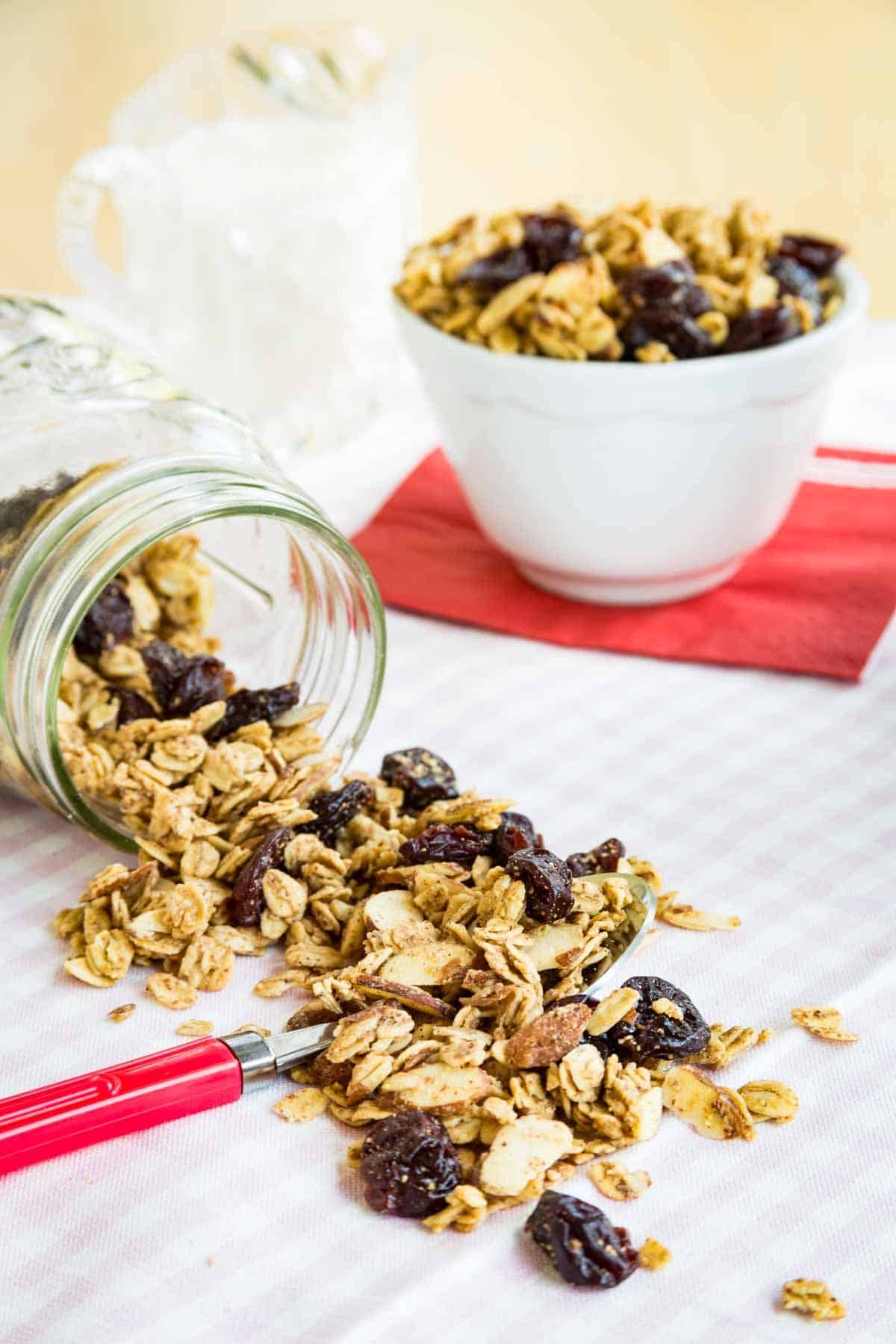 Almond granola spills out of a jar with a bowl of granola behind it.